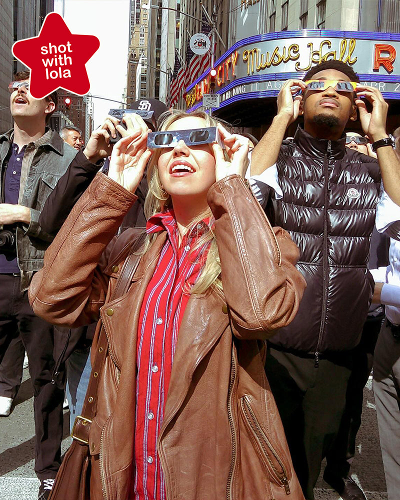 Photo taken on a Lola camera: woman in a leather jacket wearing eclipse glasses in a city crowd